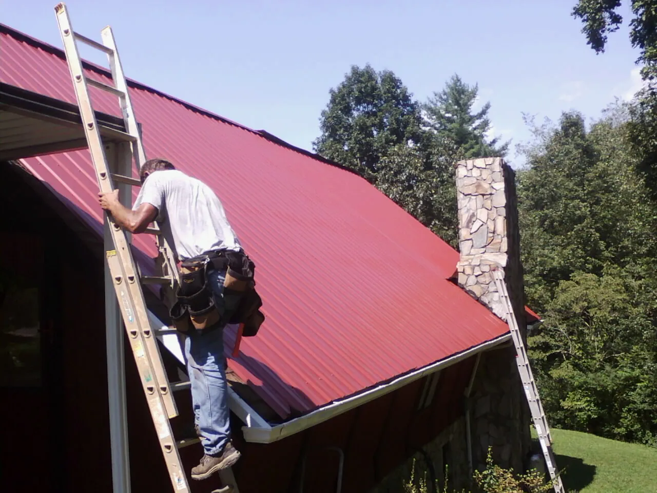Roofer on a ladder beside a red metal roof and stone chimney — Soffit & Fascia Repair by Roofing Brothers in Mango